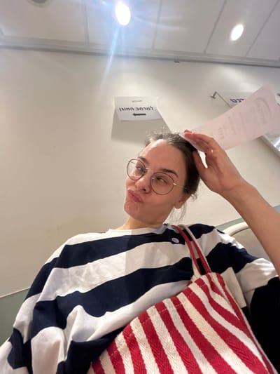 A young woman with blonde hair pulled back is taking a selfie while sitting indoors. She is wearing a navy and white striped long-sleeved shirt and round glasses. She is puckering her lips and holding a piece of paper above her head with her right hand. A red and white striped tote bag is resting on her lap, with one strap visible over her shoulder. In the background, a white wall is visible with a sign written in Hebrew that reads "למרחב המזגן" which translates to "To the air conditioning area." There are also fluorescent lights visible on the ceiling. The setting appears to be a waiting area or common space, possibly in the city of Rehovot, Israel, given the Hebrew signage.