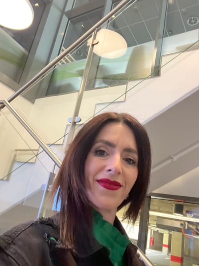 The image is a self-portrait (selfie) of a woman, presented rotated 90 degrees counter-clockwise. The woman, who is the central subject, has medium-length dark brown hair, dark eyebrows, and dark eyes. She is wearing bold red lipstick and has a light complexion, exhibiting a slight, pleasant smile. Her attire includes a green garment visible around her neck area, worn underneath a dark, possibly black or dark grey, jacket with a textured or worn collar.
The setting appears to be indoors within a modern building, characterized by contemporary architecture. The background features extensive use of glass panels, some reflecting light, and sleek metal railings, suggesting a staircase or an architectural divider. White walls and ceiling structures, including what looks like an air vent and recessed lighting, are also visible. The clean lines and bright, indirect lighting contribute to a contemporary aesthetic.
The woman is looking directly at the camera, capturing herself in the foreground while the modern interior forms the background. The overall impression is one of an individual in a bright, modern public or commercial space.
In the background, visible through a glass partition, there is a sign with the French word "SORTIE" (Exit) printed on it in capital letters. Other indistinct details and machinery-like elements can be vaguely seen through distant windows or glass walls. The location context provided is Mensdorf, Luxembourg, which aligns with the modern European architectural style seen.