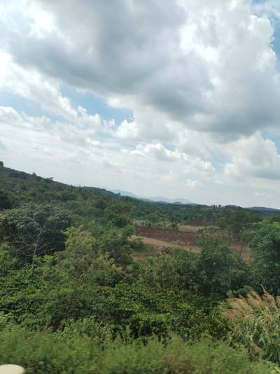 The image depicts a landscape in Thôn 8, Vietnam, under a cloudy sky. Lush green vegetation dominates the foreground and mid-ground, with trees and shrubs covering the terrain. In the distance, a cultivated field of reddish-brown earth is visible, indicating agricultural activity. Further beyond, rolling hills or mountains can be seen on the horizon.