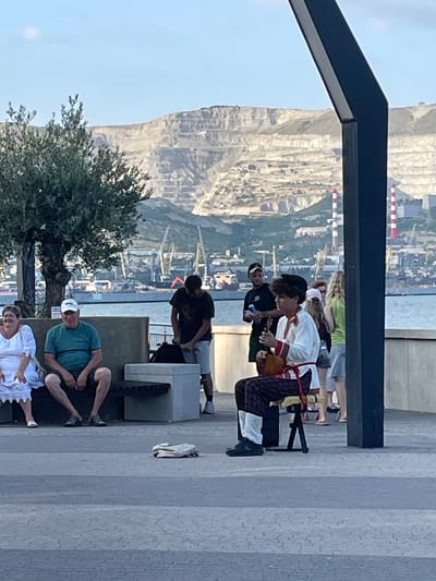 The image captures a group of people gathered outdoors, with a scenic backdrop of a rocky mountainside, a body of water, and a distant industrial port. The scene appears to be set on a paved promenade, possibly a boardwalk or pier, given the presence of a dark, angular structure overhead, suggesting a modern architectural element.

In the foreground, three individuals are seated. One person, wearing a green t-shirt and a white cap, is sitting with their legs crossed, looking towards the camera. Next to them, another person in a white outfit is partially visible, also seated. A third person, unseen, is likely sitting beside them.

Further back, a group of people are standing and interacting. One individual, dressed in a white shirt with red trim and patterned trousers, is seated on a small stool, holding a long, thin object that could be a musical instrument or prop. Another person is leaning over them, and a few other figures are standing around, some appearing to be onlookers. The clothing suggests a casual setting, possibly a warm day.

The background reveals a vast, layered mountain range with visible striations. Below the mountain, a complex of industrial structures, including cranes and smokestacks, lines the waterfront, indicating a working port. The sky is clear and light, suggesting daytime, likely late afternoon or early evening due to the quality of light. The overall mood seems relaxed and informal, with people enjoying the view and each other's company. There is no visible text in the image. The location context is Ivanovskaya, Russia.