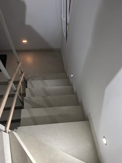 The image shows a set of stairs in a residential building in Limenaria, Greece. The stairs are made of light-colored stone and are illuminated by small, recessed light fixtures on the wall. The handrail is made of metal and wood. The floor is tiled. The scene is empty of people. The image captures the staircase from the bottom, looking up towards the top. The stairs are set against a white wall and are part of an interior space. There is a window on the right side of the image, looking out towards an unknown exterior. The image is taken at a slightly downward angle, showing the staircase from the bottom. The image is well lit and the colors are muted. The scene conveys a sense of simplicity and peace.