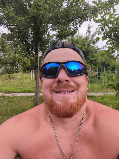 A medium shot selfie shows a man with a ginger beard and wearing sunglasses and a baseball cap, standing outdoors. The man is shirtless, revealing a silver chain around his neck. The background features trees, grass, and a fence, suggesting a rural or suburban setting. The scene is likely in Perliavka, Ukraine, given the context.
