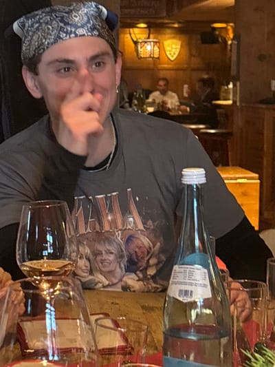 The image displays an indoor scene within a restaurant or tavern in St. Christoph, Austria. A young male individual, centrally positioned in the foreground, is seated at a light-colored wooden table. He has dark hair covered by a blue and white patterned bandana, wears a dark grey t-shirt with a "MAMMA MIA!" graphic featuring three female faces, and a black long-sleeved shirt underneath. A silver chain necklace and a small hoop earring are visible. His right hand is raised, with fingers obscuring his nose and mouth, while his facial expression suggests amusement. On the table, a wine glass holding rosé liquid is prominent on the left, alongside a clear glass bottle with a white screw cap, partially filled with water, and bearing a label. Additional empty glassware and red napkins are also present. The background features a warmly lit interior with wooden wall paneling, decorative wall sconces, and two crest-like wall ornaments. Several blurred figures, identified as other patrons, are visible in the distance at separate tables.