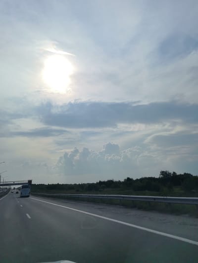 The image depicts a highway scene with a bright sun partially obscured by clouds. A white bus is visible driving on the road, along with other vehicles in the distance. The road has clear white lane markings. A metal guardrail runs along the side of the highway, separating it from a grassy area and a line of trees in the background. There are also streetlights visible on the left side of the image. The sky is partly cloudy with dramatic cloud formations. The scene appears to be set in a rural or suburban area outside of a city, possibly near Timiryazevsky, Russia, given the context.