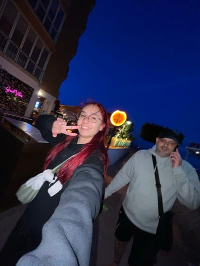 The image depicts two individuals in an urban environment at dusk or early night in Magaluf, Spain. In the foreground, a young adult female with long red hair, glasses, and a dark long-sleeved top is taking a selfie. She is smiling and making a peace sign with her right hand, her arm extended towards the camera. A light-colored cross-body bag with a white garment tied to it is visible on her left side. To her right and slightly behind her, an adult male is walking. He wears a dark cap or beanie, glasses, a light grey long-sleeved hoodie, dark shorts, and a black cross-body bag. He is holding a dark-colored smartphone to his right ear, indicating he is on a call.
The background features a multi-story building on the left. Below it, a commercial establishment displays a neon sign reading "Good Vibes" on a wall adorned with foliage. Further into the mid-ground, an illuminated orange circular sign with an abstract pattern is visible. The sky is a deep blue, characteristic of twilight.