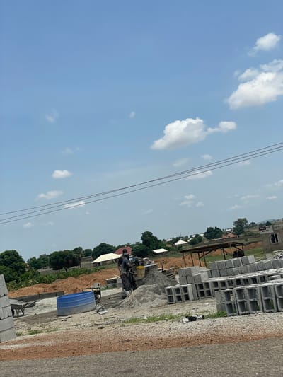 The image depicts an outdoor scene in Kwali, Nigeria. The foreground shows a stack of grey concrete blocks, likely construction materials. A grey asphalt road runs alongside the blocks. Further in the background is construction equipment, piles of gravel, and a blue container. Beyond this, trees and low-lying buildings are visible under a sky with scattered clouds and power lines. There is no indication of any people. The scene implies ongoing construction or industrial activity.