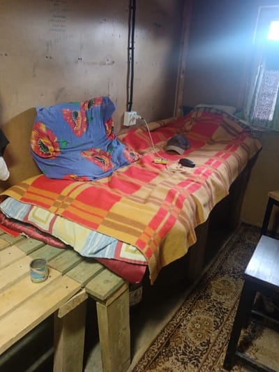 The scene is a sparsely furnished room in Яльгелево, Russia. A bed constructed from wooden planks is the central feature, covered with layers of bedding including a floral-patterned pillow, a plaid blanket, and multiple thin mattresses. Personal items such as a baseball cap, charging cable, and what appears to be a pair of glasses rest on the bed. Adjacent to the bed is a makeshift table made of similar wooden planks, upon which sits an open tin can. A rug with a floral pattern covers part of the floor. The walls are plain and have some handwritten notes on them. A small window allows light into the room, creating a bright spot. An electrical outlet and wiring are visible on the wall behind the bed. A simple wooden chair stands near the bed.