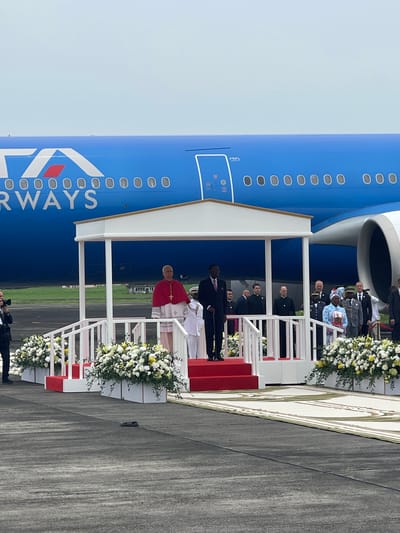 A formal outdoor ceremony is depicted at an airport tarmac under a clear, bright sky. The central figure is a man, clearly identifiable as Pope Francis by his white cassock, red mozzetta, and white zucchetto, standing at the foot of a grand, red-carpeted aircraft stairway. He appears to be greeting or being greeted by officials.

The stairway is elaborate, featuring white railings adorned with abundant white and yellow floral arrangements in planters, and is topped by a white canopy. Several people attend the event; these include men in dark suits, military uniforms, and one individual wearing traditional attire, positioned on and around the stairs.

To the left of the stairs, a large blue passenger airplane is prominent, displaying the "ITA AIRWAYS" logo and text along its fuselage, along with several windows. The "ITA" logo is also visible near one of the aircraft's doors. A person holding a camera, likely a photographer, is visible in the foreground, capturing the event. The scene conveys a sense of solemnity and official ceremony, characteristic of a high-profile arrival or departure, consistent with an event in Malabo, Equatorial Guinea.
