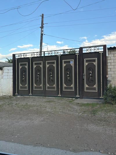 This image depicts a dark brown metal gate with ornate scrollwork and decorative trim. The gate appears to be a double gate, with four panels on each side. The panels have rectangular insets with a silver border, and within each inset, there is a black scroll design with small silver embellishments. A silver handle is visible on the upper section of the left gate. One of the panels on the left gate has a blue sign with white numbers "201". The gate is set against a backdrop of a bright blue sky with scattered white clouds. To the left of the gate, a wooden utility pole with several wires is visible. The ground in front of the gate is a rough, gray, gravelly surface with some patches of green grass. In the background, a brick wall can be seen. The lighting suggests it is daytime, likely sunny. The scene appears to be an outdoor setting, possibly an entrance to a property in a rural or suburban area. There are no people or animals visible in the image.