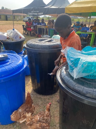 The image displays an outdoor scene in Itobe, Nigeria, specifically at the CWO Itobe Parish, indicated by a banner on a black tent. In the immediate foreground, a young male, appearing to be a child, with dark skin and an orange collared shirt, stands facing downwards, handling small clear plastic packets with both hands between two large black plastic bins. To his left, a large blue plastic bin with a lid is visible, along with another black bin filled with white plastic containers. On top of the black bin to the child's right, a light blue plastic basket covered in clear plastic is present. Two brown hens stand on the dry, light-colored dirt ground in the lower-left portion of the frame. In the midground, under a black tent and a yellow tent, multiple adults are seated on black plastic chairs at green plastic tables. A woman in a red dress is visible under the black tent, and trays of eggs are discernable on tables. In the background, a white SUV is parked near a building, and several individuals are walking across an open, dusty field under a clear sky.