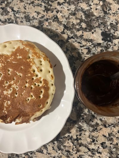 A close-up, overhead shot displays a single pancake on a white, scalloped plate positioned on a speckled granite countertop. To the right of the pancake, a small glass jar, partially filled with a dark, viscous substance, sits on the countertop. A spoon is visible inside the jar, stirring the contents. The pancake is golden brown on top with several small bubbles, suggesting it has recently been cooked. The overall setting appears to be a kitchen or dining area. The image does not contain information to determine its location in Figueres, Spain.