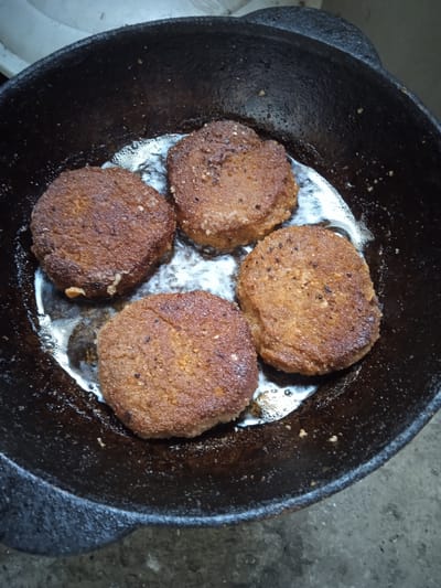 Four round, breaded patties are being fried in a dark, cast-iron skillet. The patties are a golden-brown color and appear to be seasoned with small dark specks. Bubbles of cooking oil surround the patties, indicating they are actively being fried. The skillet is set on a rough, gray surface, likely concrete. The background is out of focus, showing a light-colored, possibly ceramic surface. The scene appears to be an outdoor setting, possibly a yard or patio, in the city of Aznvadzor, Armenia, given the rustic nature of the cooking setup. There are no people visible in the image.