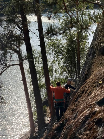 Two individuals are rock climbing on a steep, rocky incline near a body of water, likely a lake or river, in Ruch'i, Russia. The person in the foreground, wearing an orange long-sleeved shirt and a climbing harness, is facing away from the camera and appears to be preparing to climb. The second individual, wearing a helmet and a dark long-sleeved shirt, is positioned slightly behind and to the right, also equipped with a climbing harness. Both individuals are attached to climbing ropes. The rocky surface is textured and appears to be covered in moss and small vegetation in places. The background features a sun-dappled body of water with sparkling reflections, framed by the trunks of tall trees and green foliage. The overall lighting suggests a bright, sunny day.