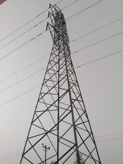 The image displays a large, imposing metal lattice-work electricity pylon (transmission tower) viewed from a low angle against a uniformly bright, overcast sky. The pylon, constructed from numerous intersecting metal beams, dominates the center of the frame, extending from the bottom edge and tapering upwards beyond the top. Multiple high-tension power lines radiate horizontally from the upper sections of the pylon, stretching across the gray sky.
In the lower-left corner, partially obscured by the base of the main pylon, a smaller, darker utility pole is visible, equipped with insulators and wires, likely for local power distribution. The very bottom edge of the image shows sparse, dark foliage, suggesting the presence of trees or bushes at ground level.
The sky is a consistent light gray, indicative of an overcast day or perhaps a hazy atmosphere, providing diffused lighting with no direct shadows. There is no discernible activity taking place, nor are there any people present. The scene is a static depiction of electrical power infrastructure. No visible text is present in the photograph. The overall impression is one of scale and industrial purpose.
