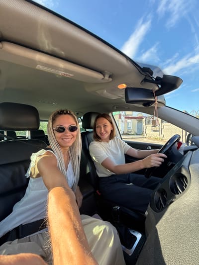 Two women are inside a car, with one woman in the driver's seat and the other in the passenger seat, both looking at the camera and smiling. The driver, wearing a white t-shirt and dark pants, has short brown hair and is holding the steering wheel. The passenger, with long blonde braided hair and sunglasses, is wearing a white dress. The car's interior, including the seats, dashboard, and rearview mirror, is visible. Outside the car, a blue sky with clouds and some buildings can be seen, suggesting an outdoor urban or suburban setting, potentially in the city of Esbol Rural District, Kazakhstan. The lighting indicates it is daytime.