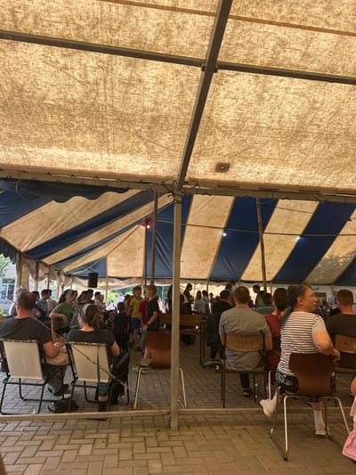 The image depicts an outdoor gathering under a large tent with a beige and blue striped fabric roof supported by metal poles. The ground is paved with light brown interlocking bricks. A diverse group of people, including adults and children, are seated and standing, facing towards the left side of the frame where an unseen event is likely taking place. Some individuals are looking towards the front, suggesting they are engaged in watching or listening to something. Several rows of chairs are visible, with some occupied by attendees. A black speaker is mounted on a pole. The environment appears to be an open-air event space, possibly a park or community gathering area, located in or near Завалочыцкі сельскі Савет, Belarus. The lighting suggests it is daytime, with sunlight filtering through the tent fabric, creating diffused illumination.