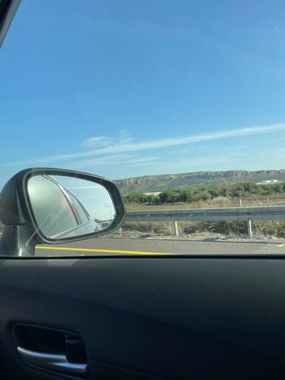 The image displays a roadside view from within a vehicle, likely on a highway near Jisr az-Zarqa, Israel, under a clear, bright blue sky. In the foreground, the dark gray/black interior door panel of the vehicle is visible, including a chrome door handle on the lower left. A bright yellow painted line delineates the edge of an asphalt road surface outside the vehicle. To the left, a dark-framed side-view mirror reflects a blurred red vehicle and another darker vehicle behind it, along with partially visible text "OBJECTS IN MIRROR ARE CLOSER THAN THEY APPEAR." Immediately beyond the road, a silver-colored metal guardrail stretches horizontally. The area behind the guardrail consists of dry, sparse vegetation, transitioning to a denser band of green and brown foliage. In the far background, a long, rugged, light brown cliff face or ridge dominates the horizon, with indistinct light-colored structures visible near its base on the right. The sky above is predominantly clear, exhibiting a few wispy white clouds.