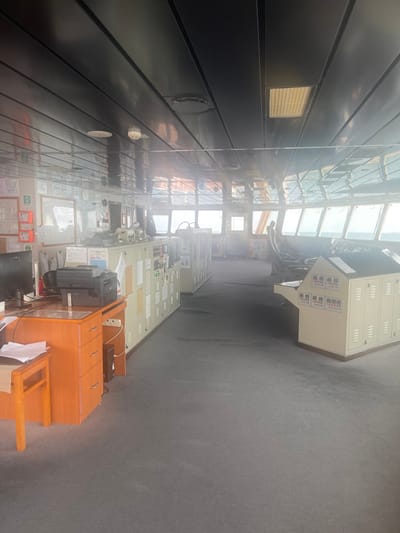 The image displays the interior of a vessel's bridge, potentially located near Orimedu, Nigeria. The space features a dark gray carpeted floor and a dark, reflective paneled ceiling with integrated circular and rectangular light fixtures and a central smoke detector. On the left, a light brown wooden desk with three drawers supports a black computer monitor and a black multi-function printer. Various papers are scattered on the desk and an adjacent wooden stool. Along the port side wall, a series of light-colored control consoles or cabinets are equipped with numerous panels, switches, and affixed documentation. Wall-mounted emergency equipment and diagrams are visible above and behind this console row. Towards the bow, another arrangement of light-colored control consoles, some featuring slanted top panels with indicators, fronts a panoramic array of large multi-pane windows. These windows offer a bright, hazy view of open water and sky; distant, indistinct elements are visible through the haze. An unidentifiable human silhouette is faintly visible in the far background, positioned between two forward control consoles. The scene appears static, with no active personnel or interactions observed.