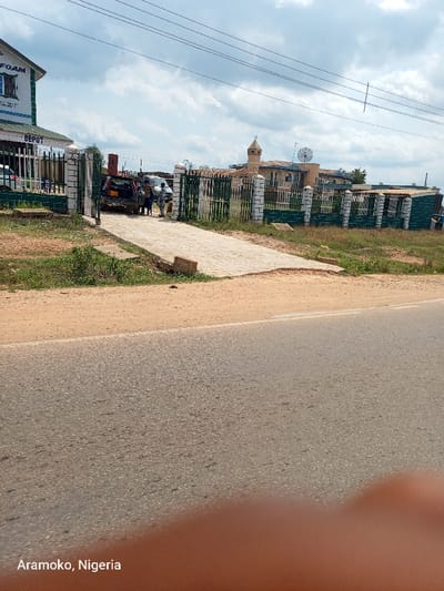 The image shows a paved driveway leading to a gated compound in Okinni, Nigeria. The driveway is lined with green vegetation and has a cracked surface. Three people can be seen walking towards a black car parked inside the compound. Two other vehicles are visible behind the car, one of which is a white sedan. The gate is made of green metal and has a concrete base. The building beyond the gate appears to be a mosque with a minaret and a satellite dish on the roof. The sky is partly cloudy. There are several power lines running across the image. The compound is located at a junction with an asphalt road, with a light brown dirt shoulder. There are a few white lines marking the edge of the asphalt. The photo was taken from a car. The scene is typical of many residential areas in Nigeria.