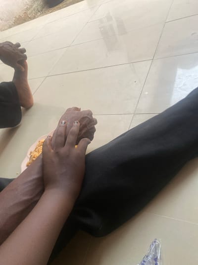 The image shows a close-up view of two people's arms and hands, with a plate of food in the foreground. One person is wearing black pants and their arm is visible from the elbow down, resting on the floor. Another person's hands are intertwined with the first person's. One hand has dark nail polish on its fingernails. A small, pink plate with food, possibly rice or noodles, is visible between their hands. The floor appears to be tiled with light-colored square tiles. The scene is indoors, possibly in a home or a public space. The lighting suggests it could be daytime. There are no discernible location cues beyond the tiled floor and the general suggestion of an indoor setting. The overall impression is one of intimacy or comfort, with the intertwined hands and shared meal.