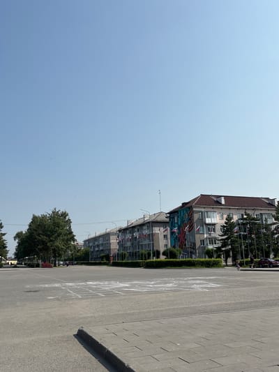 The image depicts a wide, paved town square in Belovo, Russia, on a clear, sunny day. In the foreground, a hopscotch grid is drawn in chalk on the asphalt. To the left, a large, leafy tree stands prominently. Behind the tree and further into the scene are low-rise apartment buildings lining the square. The buildings are adorned with flags, and one building features a mural. Several evergreen trees are planted in front of the buildings. A few people are visible walking in the distance, and a red car is parked on the right side of the square. The overall impression is of a quiet, ordinary day in a public urban space.