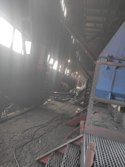 The image depicts the interior of an industrial facility, likely a workshop or factory, with a raw, unfinished aesthetic. Natural light streams in from large windows on the left, creating bright areas and casting long shadows. The floor is covered in a coarse, dusty material, possibly concrete or dirt, with various cables and wires strewn about. In the mid-ground, a person wearing an orange hard hat is bent over, seemingly engaged in some form of work or repair. To the right, a large blue metal structure, possibly part of machinery or equipment, dominates the scene. The background reveals a high ceiling with exposed beams and corrugated roofing. The overall atmosphere is one of active industrial work, with elements of construction or maintenance visible. The location is identified as the city of Kyzyl, Kazakhstan.