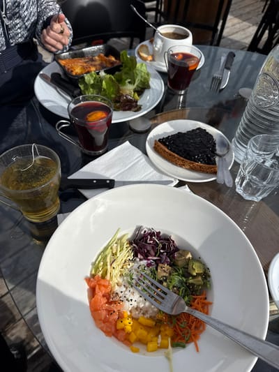 The scene depicts an outdoor dining setup on a glass-top table in Saint-Martin-de-Belleville, France. A person's hands, wearing a patterned long-sleeved top and a ring on the right ring finger, are visible in the upper left, holding cutlery over a meal. In the foreground, a large white bowl contains a poke-style dish with segmented portions of white rice, diced salmon, diced yellow mango, shredded carrots, shredded red cabbage, shredded green vegetables, and avocado cubes, topped with sesame seeds. A silver fork rests within this bowl. To its left, a clear glass mug holds green tea with a visible tea bag. A white paper napkin and a black-handled knife are placed between these items. Further back on a white plate, a black baking dish contains a gratin alongside a green leaf salad. Two clear glass mugs, each with a handle, contain a dark red beverage garnished with an orange slice. A small white plate presents a slice of dark berry tart with two silver spoons. A white cup containing a dark beverage and a small piece of bread on a saucer is also visible. A large, textured glass water bottle and multiple empty clear drinking glasses are positioned on the table's right side.