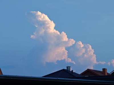 This image captures a twilight sky over Dörpen, Germany, with dramatic cumulus clouds illuminated by the setting sun. The clouds are the dominant feature in the upper portion of the frame, displaying vibrant shades of white and pink against a deep blue sky. Below the clouds, the dark silhouettes of rooftops are visible, punctuated by chimneys. One chimney with a silver cap stands out on a dark-tiled roof in the foreground. The lower edge of the image is obscured by a dark, horizontal structure, possibly a building or overhang. The scene appears serene, with no people or animals present, focusing entirely on the interplay of light, cloud formations, and urban architecture.
