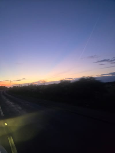 The image depicts a twilight scene on a rural road, possibly in the vicinity of Lichfield, United Kingdom. The sky transitions from a deep blue overhead to shades of purple, pink, and orange near the horizon, suggesting either dawn or dusk. Faint clouds are visible, and a distinct contrail stretches across the upper portion of the sky. A line of trees and foliage forms a dark silhouette along the right side of the road. A single lamppost stands erect near the left side of the road, and the road itself appears wet, reflecting the ambient light. The perspective suggests the photo was taken from a moving vehicle, as indicated by the motion blur in the foreground, particularly the illuminated headlights casting a yellow glow on the road surface and the blurred outline of what appears to be a vehicle's interior on the bottom left. The overall impression is one of a journey on a quiet road during the transitionary hours of daylight.
