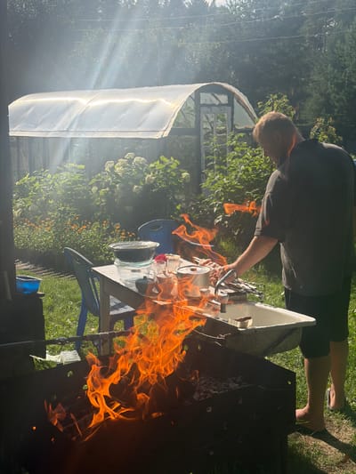 In an outdoor setting with a greenhouse in the background, a man is standing and preparing food over an open fire. He is wearing a dark t-shirt and dark shorts. A barbecue grill with flames is in the foreground. A table is set up with various items, including bowls, glasses, and utensils. The surrounding environment is grassy with trees and bushes. The scene appears to be in a residential area, possibly a dacha or a backyard, in Белая Холуница, Russia.