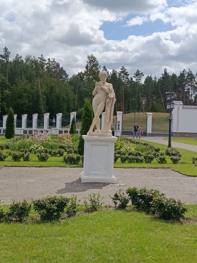 A marble statue of a nude male figure stands on a marble pedestal in the foreground of a formal garden. The statue is positioned to the right of center. The garden features a gravel path, neatly trimmed green bushes, and flower beds with pink and white flowers. A white fence with black wrought-iron accents and regularly spaced pillars encloses the garden. Beyond the fence, a dense line of green trees forms the background, with a cloudy sky above. In the middle ground, two people are walking on a paved path near a large decorative gate. A black lamppost is visible to the right of the gate. The scene is set in the park of the Kosava Castle, Kosava, Belarus, under a partly cloudy sky.