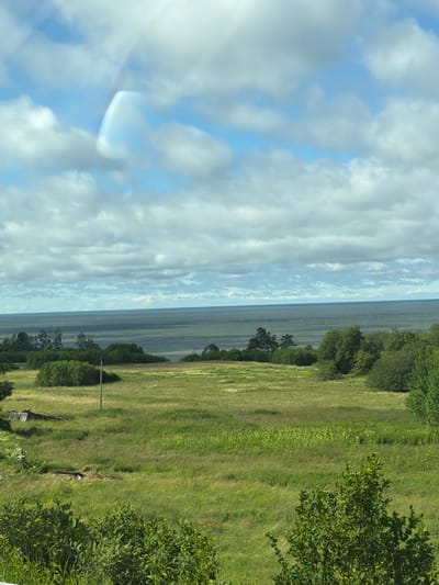 The image captures a scenic view of a coastal landscape in Aseri, Estonia, during what appears to be a clear day with a mix of sun and clouds. The scene is divided into three distinct horizontal sections.
The left third of the image is dominated by a vast expanse of sky filled with fluffy white clouds against a bright blue backdrop. Sunlight streaks through the clouds, creating a luminous and ethereal effect.
The middle section depicts a calm body of water, likely the Baltic Sea, stretching towards the horizon. The water's surface is smooth, reflecting the sky and appearing a deep, muted blue. Along the coastline, a line of trees marks the transition between the water and the land.
The right third of the image showcases a lush, green grassy field. The grass is a vibrant green, with some areas appearing slightly lighter due to the sunlight. Scattered throughout the field are small yellow wildflowers. A few trees are visible, their dense foliage adding depth to the scene. In the distance, a single wooden post stands in the field, hinting at the presence of human activity or past structures. The overall impression is one of natural beauty and tranquility.