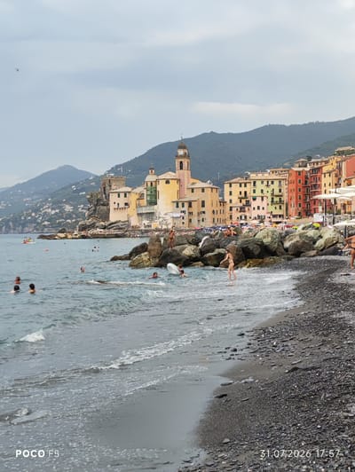 The image shows a view of the beach and town of Camogli, Italy. In the foreground, the choppy blue sea is in motion with small waves breaking on a dark, pebbly beach. Several people are visible in the water, some swimming, others standing. On the right side of the image, a few large rocks are scattered along the shoreline, and a person is standing on one of them. Further back, the beach is lined with beach umbrellas and lounge chairs. Behind the beach, colorful buildings rise up the hillside, with a prominent church tower featuring a clock. Mountains form the backdrop to the town, and the sky is overcast. A small red helicopter is visible in the sky far in the distance. The bottom of the image has text overlays indicating "POCO F5" and "31.07.2026 17:57".