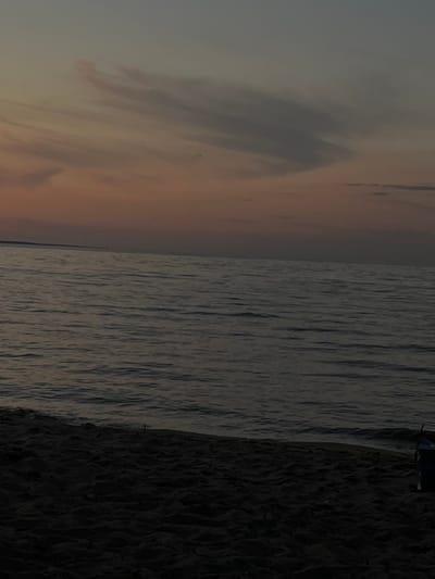 The image depicts a natural scene at dusk, likely a beach or shoreline. The sky is a gradient of dusky oranges and grays, with wispy clouds stretching across the upper portion of the frame. Below the sky is a body of water, appearing to be a lake or sea, with gentle ripples on its surface. In the foreground, dark sand forms a beach. A small, dark object, possibly a piece of driftwood or debris, is visible on the sand near the waterline. To the far right, a dark blue object, potentially a bag or container, is partially visible on the sand. The lighting suggests it is either sunrise or sunset, with the overall mood being serene and quiet. There are no people or animals visible in the scene. The provided crops highlight the sky, the horizon line where the sky meets the water, the water itself, and the sandy foreground.