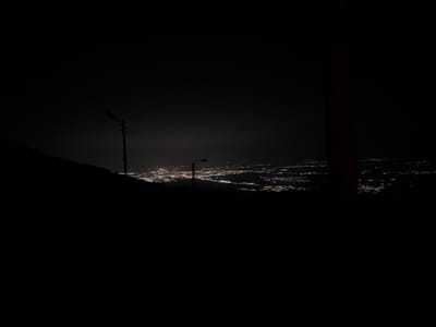 The image displays a nighttime, elevated view overlooking the city of Yavrovo, Bulgaria. A dark, silhouetted hillside occupies the lower left portion of the frame. Two dark utility poles, each equipped with a street light fixture, are visible in the midground, silhouetted against the distant urban lights. The city of Yavrovo in the background presents a wide expanse of numerous white and yellow light clusters, indicating illuminated buildings and infrastructure. The night sky is dark, exhibiting a subtle atmospheric glow directly above the city lights. A dark, vertical object is partially visible along the extreme right edge of the frame. No individuals or direct actions are discernible.