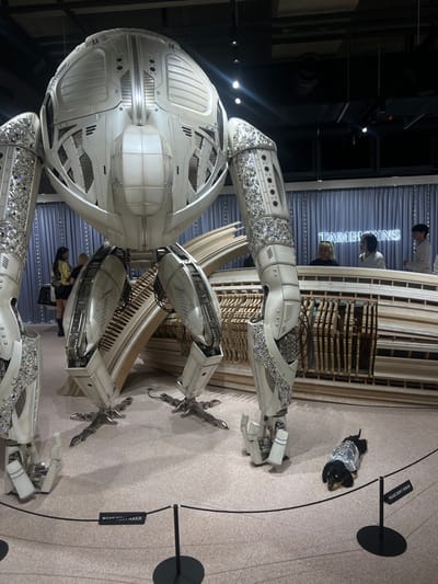 An indoor exhibition space in Seongdong-gu, South Korea, features a large, intricately designed mechanical creature sculpture positioned centrally. The sculpture is predominantly white and silver, presenting a bulbous central body with detailed paneling and perforated sections. Six robust, articulated limbs extend from the main body; four function as support legs, terminating in multi-clawed feet on the light-colored floor, while two additional arm-like structures extend forward. Sections of all six limbs are heavily adorned with numerous small, sparkling, crystal-like embellishments.
In the immediate foreground, a black and tan Dachshund, wearing a shimmering silver jacket, stands on the floor behind a low black rope barrier. A small black sign attached to the barrier explicitly states, "PLEASE DON'T TOUCH." In the mid-ground, behind the sculpture, a large, curvilinear structure constructed from multiple parallel light-colored wooden slats spans horizontally. Behind this wooden element, a light blue/grey textured wall serves as a backdrop, featuring a prominent backlit neon sign that reads "TAMBURINS." Several individuals are visible in the background and mid-ground, observing the scene; one person in a yellow top and black skirt is partially visible on the left, while others are further back near the "TAMBURINS" sign. Bright overhead lighting illuminates the entire display.