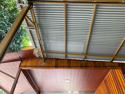 The image shows a view looking up at the underside of a covered structure, possibly a porch or an outdoor area. The ceiling consists of two distinct levels: a lower ceiling made of polished wooden planks and an upper ceiling constructed from corrugated metal sheets supported by yellow metal beams. A white security camera is mounted on one of the yellow beams, overlooking the area. To the left, lush green foliage from a tree is visible, suggesting an outdoor or semi-outdoor setting. A decorative spherical light fixture made of woven material hangs from one of the beams. The overall scene is one of architectural elements and outdoor integration, with no people or specific actions depicted. The location is identified as Aluepiet, Indonesia.