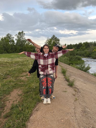 The image shows three people outdoors. A person wearing a baseball cap is standing behind a younger person wearing a red and white plaid shirt, which is covering another person. The three people are lined up and have their arms extended outward, resembling the shape of an airplane. They are standing on a grassy area next to a concrete structure. A river and wooded area are visible in the background. The sky is cloudy. The location appears to be in a park near a river in Nizhnii Vishur, Russia.