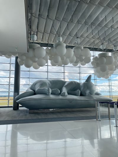 The image depicts the interior of a spacious terminal hall at Paris Charles de Gaulle Airport, Terminal 2E, located in Roissy-en-France, adjacent to Mitry-Mory. The primary subject is a large, light grey sculpture of a cat sleeping on its side atop a rectangular pillow, positioned centrally in the hall. Inscriptions "L'ESPRIT DU VENT" and "A. GILLAUD," along with Japanese text, are visible on the pillow. The ceiling features segmented, light grey paneling, from which numerous clusters of spherical, opaque white light fixtures, resembling bubbles, are suspended. A vast glass curtain wall forms the background, providing an unobstructed view of an airfield with green ground cover and distant infrastructure under a partly cloudy sky. The floor is composed of highly polished, light-colored tiles, exhibiting prominent reflections of the sculpture and overhead lighting. A dark grey cylindrical support pillar is visible on the far left, and a metal stanchion barrier is partially visible on the far right. No individuals are present in the scene.