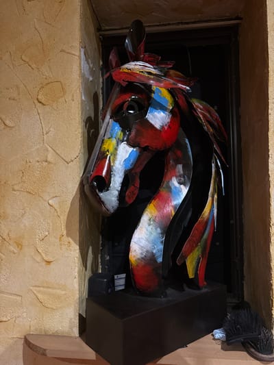 A vibrant, abstract metal sculpture resembling a horse head rests inside a fireplace alcove. Bold strokes of red, white, yellow, and blue paint adorn its metallic form. The sculpture's angular shapes mimic equine features. A black fireplace insert and hearth provide a contrasting background. A dusting brush is visible near the top of the frame. The tan fireplace surround has a textured surface.
