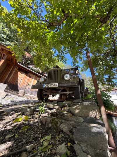 The image captures a low-angle view of a vintage olive-green military truck parked on a rocky incline in Shiroka Laka, Bulgaria. The truck, with a license plate reading "CM 2015 CM," is partially obscured by the lush green canopy of a tree overhead, which casts dappled shadows on the scene. To the left of the truck, a rustic wooden structure with a sloping roof is visible. The ground is a rough terrain of stones, dirt, and fallen leaves. In the background, a tall, rusty metal pole stands vertically to the right of the truck, and hints of houses and trees suggest a rural or mountainous setting. The sky is clear blue.