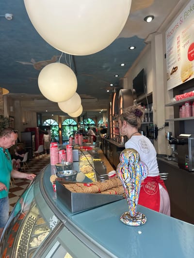 The image depicts the interior of an ice cream shop in Zwolle, Netherlands. In the foreground, a man wearing a turquoise shirt and sunglasses is leaning over an ice cream display case. To the right, a woman with blonde hair tied up and wearing a white t-shirt that says "GELATO ITALIANO ORIGINALE" and a red apron is standing behind the counter. She is holding a colorful, abstract sculpture of an ice cream cone.

The shop has a counter with various ice cream cups, waffle cones, and a metallic container filled with more waffle cones. The display case is filled with different flavors of ice cream. In the background, other customers can be seen, as well as arched doorways leading to another part of the establishment. The ceiling has a painted mural of clouds and birds, and large, spherical light fixtures hang down. A price list is visible on the wall to the right, listing prices for scoops and waffle cones in Dutch. The overall atmosphere is bright and casual, typical of an ice cream parlor.