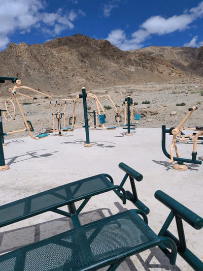 In Phyang, India, an outdoor exercise area is situated at the foot of a barren, rocky mountain range under a bright blue sky with scattered white clouds. The area is paved with a light-colored, rough surface. Several pieces of exercise equipment, primarily in a pale yellow and dark green color scheme, are arranged on the paving. These include multi-gym stations with seats and handles, some designed for chest presses or leg extensions, and a device with a large wheel. In the foreground, two dark green metal benches with perforated seating are prominent. The benches are angled towards the viewer, with one positioned in front of the other. The ground around the equipment is dry and sandy, with sparse, low-lying vegetation visible in the distance. Shadows from the equipment and benches are cast on the paved surface. The scene appears to be deserted, with no people present. The overall impression is one of an open-air fitness facility in a remote, mountainous environment.