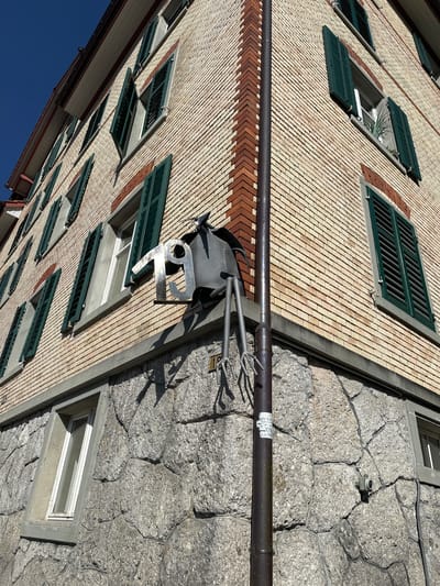 The image shows the exterior of a multi-story building, likely an apartment or office building, with a light-colored brick facade and a stone foundation. The building has several windows with dark green shutters, many of which are partially or fully open. A metal sculpture, resembling a bird or insect with a number "19" attached to it, is mounted on the brick wall near the corner of the building. A dark metal pipe runs vertically down the corner of the building, and a small security camera is visible on the stone foundation. The sky is clear and blue. The building is located at Bahnhofstrasse 19 in Wald (ZH), Switzerland, as indicated by the house number sign.
