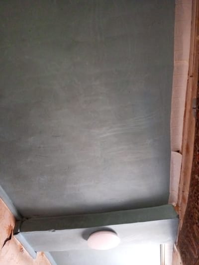 The image shows a ceiling in a room in Parabel, Russia. The ceiling is painted a light grey-blue color. There is a protruding structural element, painted the same color, that appears to partially conceal a window. A circular, white ceiling light fixture is visible on the underside of this element. Portions of a wall with floral wallpaper and a wall covered in what appears to be wallpaper with a vertical pattern are visible at the lower-left and right edges of the image, respectively. The scene is lit by daylight coming through the window. No people are visible.