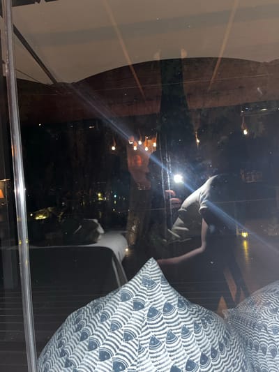 The image shows a nighttime urban scene reflected in a window. The reflection includes buildings and streetlights. A person and a fish-patterned pillow are also reflected in the window. It's not possible to confirm the city of Livingstone, Zambia from visual cues alone.