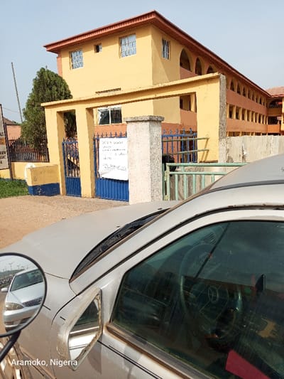 The image shows a silver car parked in front of a two-story building located in Ado Ekiti, Nigeria, at the coordinates (latitude: 7.656, longitude: 5.23). The building is yellow with an orange trim and features arched windows and a metal gate with a blue paint. A sign is posted on the wall to the left of the gate. The car is dusty, and its side window is partially rolled down, revealing the interior, including the steering wheel and the dashboard. The car's side mirror is reflecting a silver car parked in the street behind the photographer. A green bush can be seen to the left of the image, behind the building, and a small, dirt path can be seen in front of the building. The sky is clear and blue, and the sun is shining. The image is captured from the front passenger seat of the car.