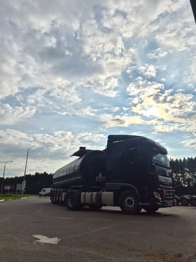 A black tanker truck is parked on a paved surface, possibly a road or parking lot, in Mozhary, Russia. The truck is facing to the right of the frame, with its front cab visible and the large cylindrical tanker body extending behind it. The sky above is partly cloudy with sunlight breaking through, casting a bright glow. In the background, a line of trees forms a dark silhouette against the sky. Other vehicles, including a white van and what appear to be spare tires or other equipment, are visible in the distance. The ground is asphalt, and a white arrow is painted on the surface, indicating a direction. The overall scene depicts a commercial vehicle in an outdoor setting under a dramatic sky.