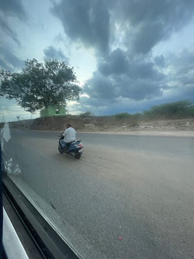 The media file shows a person on a scooter, facing away from the camera, stopped on a dusty road. The person is wearing a white shirt and blue jeans. The scooter is dark blue and white, with a license plate visible that appears to have Indian characters. The road is paved but appears to be covered in fine dust. To the left of the road, there is a barren, rocky embankment with some sparse vegetation and a large tree. In the background, a small, green building with a weathered appearance is visible. The sky is overcast with dramatic, dark clouds, suggesting a possibility of rain or stormy weather. The overall impression is that of a rural or semi-rural setting, possibly in India given the location context provided. The lighting suggests it might be late afternoon or early evening. The person appears to be resting or pausing their journey.