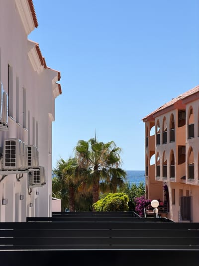 This image depicts a sunny day in Alcalà de Xivert, Spain, with a view of buildings, palm trees, and the sea. In the foreground, dark, horizontal steps dominate the lower portion of the frame. To the left, a white building with a tiled roof is partially visible, featuring several outdoor air conditioning units mounted on its exterior. To the right, a peach-colored building with arched windows and balconies is prominent. Between the buildings, lush green palm trees and vibrant purple bougainvillea are visible, with a clear blue sea and sky in the background. The scene appears to be part of a resort or residential complex.